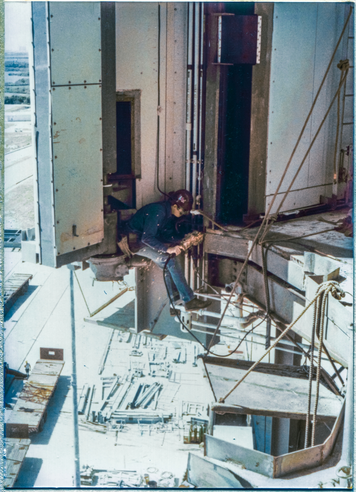 Image 059. Union Ironworker. Local 808. Working for Ivey's Steel Erectors on the Rotating Service Structure at Space Shuttle Launch Complex 39-B, Kennedy Space Center, Florida. I do not recall this man’s name, alas. No harness. This the way we did the work, back in the early 1980’s, and nobody minded, and nobody ever fell, and nobody ever even got sensibly hurt. He is grinding a weld smooth, which he just laid in to the new framing steel at elevation 135’-7” which is the Main Floor level of the Payload Changeout Room, and which abuts the top edge of the orbiter’s OMS Pods when the RSS is mated to it, and which was extensively modified to accommodate the installation of the OMS Pod Heated Purge Covers, which turned out to be a spectacularly-bad idea, both conceptually and physically. We did not like the OMS Pod Heated Purge Covers, and those ironworkers who were the most savvy regarding the ways of things like this disliked them the most. They knew what was coming. And they told those of us in the field trailers, and we told NASA, and NASA handed it to their best engineers to give it their regarded consideration… and then told us to proceed with the Covers, anyway. And just as those most-savvy ironworkers said it would, it did not end well. Photo by James MacLaren.